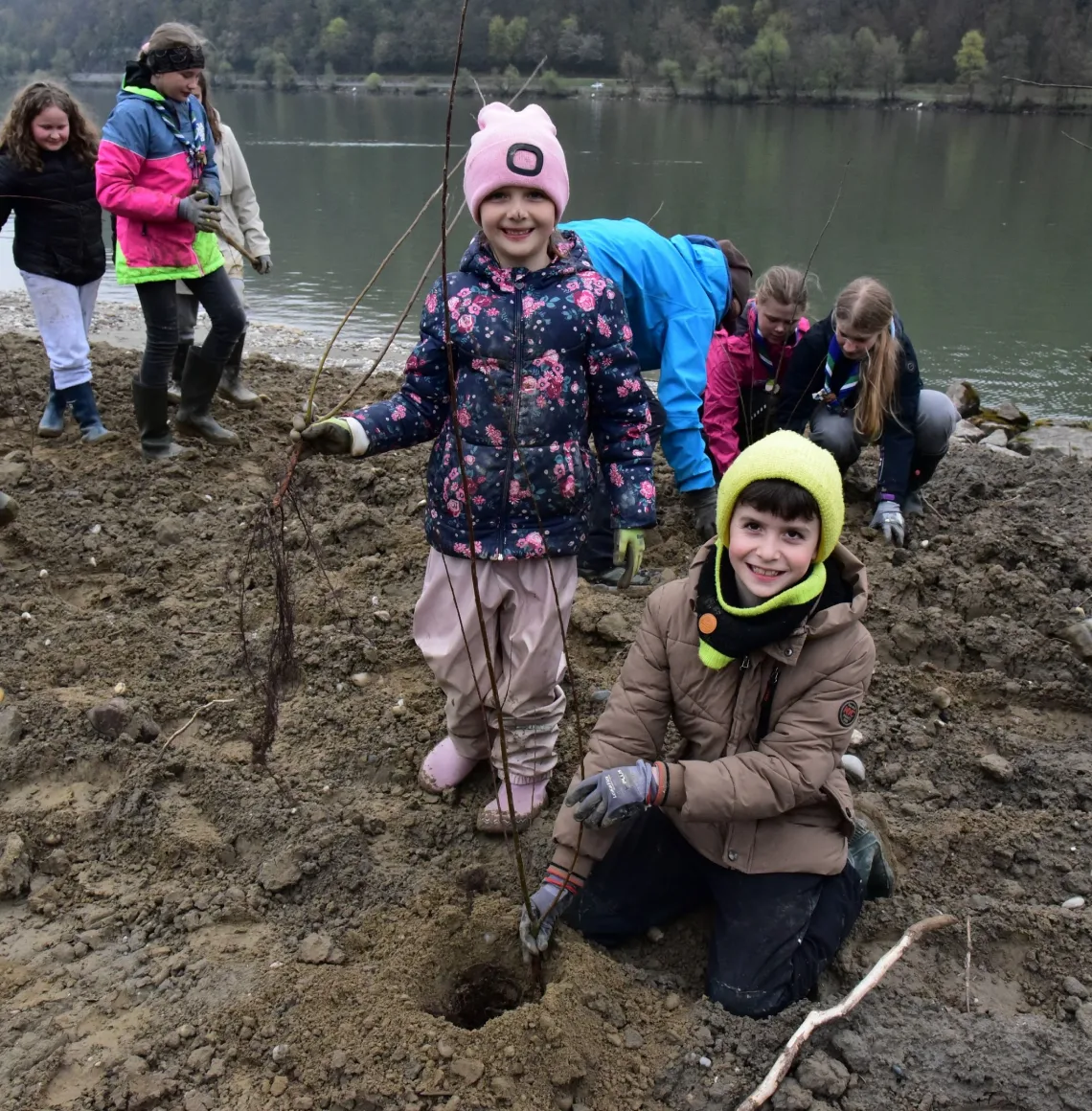 Kinder in wetterfester Kleidung pflanzen Jungbäume am Ufer eines Gewässers. Sie stehen und knien auf feuchtem Boden, viele halten Setzlinge in den Händen. Im Hintergrund ist eine bewaldete Landschaft zu sehen.