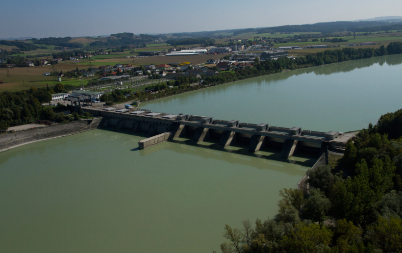 Das Bild zeigt eine Luftaufnahme des Wasserkraftwerks Schärding-Neuhaus. Im Mittelpunkt ist das Kraftwerk und die blaugrüne Donau zu sehen. Im rechten Bildrand sieht man Wald und hinter dem Kraftwerk ist die Stadt Schärding sowie die Landschaft mit strahlend blauen Himmel sichtbar.