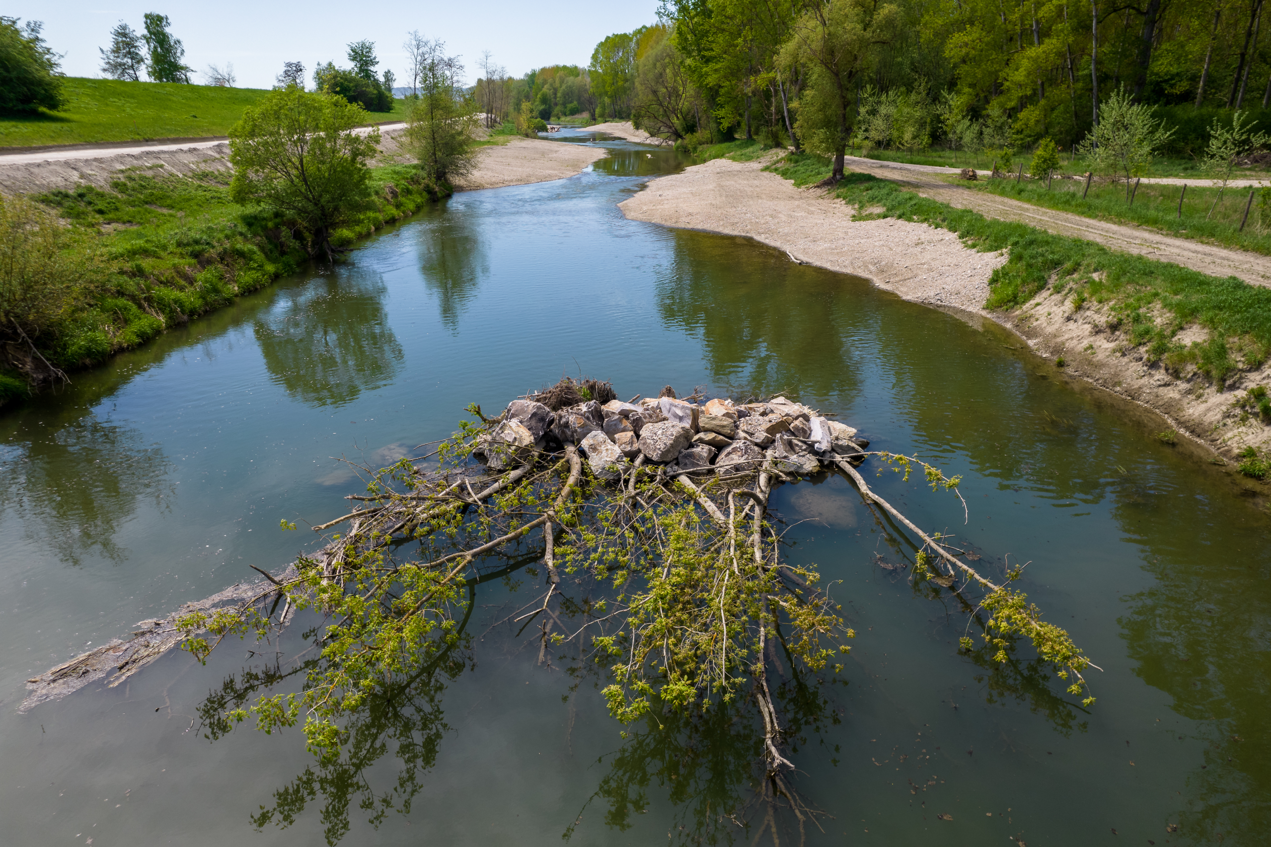 The picture shows a renaturalised site at the fish migration aid in Altenwörth. At this point in the blue-green river there is a collection of stones with plants, just like those found in nature. To the left and right of the banks you can see gravel and green areas with shrubs and trees. The blue sky shines in the upper part of the picture.